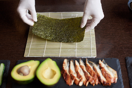 Close-up view of process of preparing rolling sushi. Chef's hands are holding sheet of nori. Avocado and eel on black stone platesの写真素材