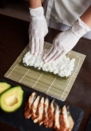 Close-up view of process of preparing rolling sushi. Nori and white rice on bamboo mat. Chef's hands touch rice. Chef starts cooking sushi. Eel and avocadoの写真素材