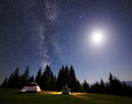 Camping night in mountain valley. Alone tourist sitting on grass at tent enjoying beautiful clear starry sky and fool bright moon over pine trees. Beauty of nature, tourism and traveling concept.の写真素材