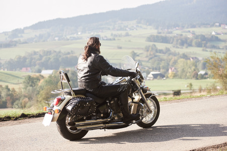 Long-haired bearded biker in sunglasses and black leather outfit driving cruiser powerful motorcycle along sunny asphalt road on bright summer day on background of green rural misty landscape.の写真素材