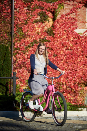 Young happy long-haired smiling blond woman in casual clothing and glasses cycling pink lady bicycle on bright warm sunny day on background of brick wall overgrown with beautiful red ivy leaves.の写真素材