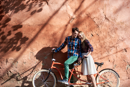 Young happy romantic tourist couple in casual clothing, bearded man and blond woman in glasses leaning together on double tandem bike on background of high red old crackled plastered wall.の写真素材