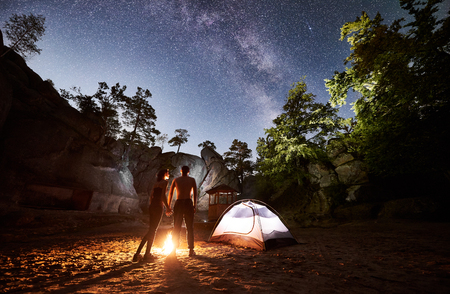 Back view silhouettes of young couple hikers man and woman having a rest beside camp, bonfire and illuminated tourist tent in mountains, enjoying beautiful night starry sky full of stars and Milky wayの写真素材