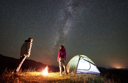Happy couple hikers having a rest at summer night camping in mountains. Young man and woman standing beside campfire, illuminated tourist tent under beautiful starry sky full of stars and Milky wayの写真素材