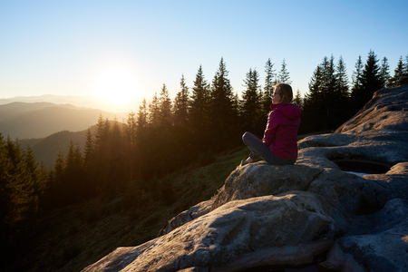 Young woman hiker sitting on big rock on the top of mountain in the evening. Female tourist enjoying incredibly beautiful sunset. On background forest, setting sun and blue skyの写真素材