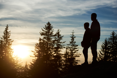 Silhouettes of young father and son standing on the top of mountain in the evening. Man and child enjoying beautiful sunset, trees on blurred background.の写真素材