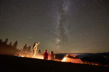 Happy friends hikers having a rest together around campfire beside camp and tourist tent at night in the mountains. Man pointing at incredibly beautiful night starry sky full of stars and Milky way.の写真素材