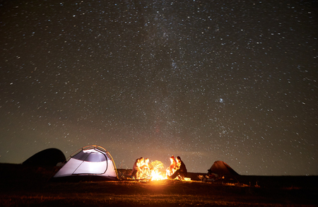 Young friends hikers having a rest together around bonfire beside camp and glowing tourist tent at night in the mountains. On background beautiful night starry sky full of stars and Milky way.の写真素材