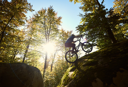 Silhouette of professional cyclist riding on back wheel on trial bicycle, sportsman making acrobatic stunt on the edge of big boulder in the forest on summer sunny day. Concept of extreme sportの写真素材