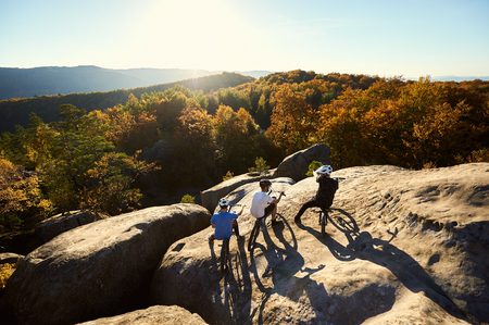 Back view of three young sportsmen with trial bicycles having a rest on the big boulder on the top of mountain. Guys enjoying beautiful view of setting sun and forest. Concept of extreme sportの写真素材