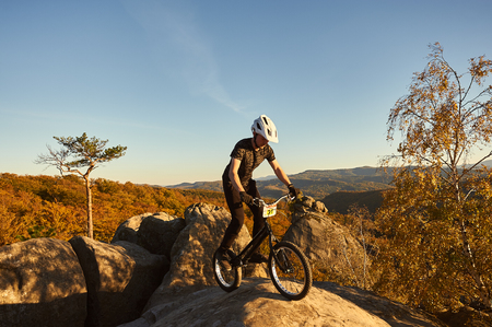 Young male cyclist balancing on trial bicycle on top of big boulder, rider making acrobatic stunt on summer evening, blue sky and forest on background. Concept of extreme sport active lifestyleの写真素材