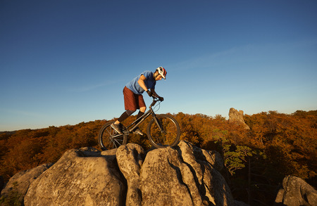 Young male cyclist balancing on trial bicycle on top of big boulder, rider making acrobatic trick on summer evening, blue sky and forest on background. Concept of extreme sport active lifestyleの写真素材