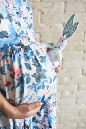 Cropped view of pregnant woman in dress standing, embracing big tummy and keeping lovely toy bunny near belly. Young cute female waiting for infant. Concept of love and pregnancy.の写真素材