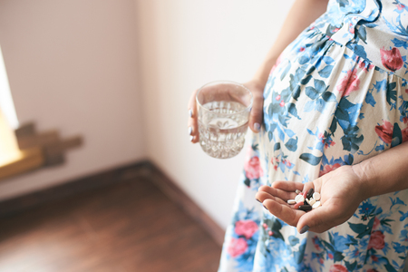 Side view of hands of pregnant woman keeping pills and glass of water. Young female in flowery dress waiting for child and taking vitamins for health of baby. Concept of anticipation and care.の写真素材