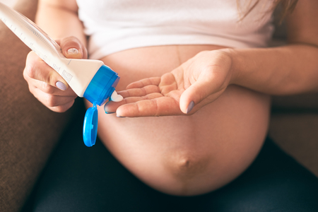 Selective focus of female hands keeping cream and applying it on big belly. Young pregnant woman sitting on sofa and caring about skin. Concept of expectation and parental love.の写真素材
