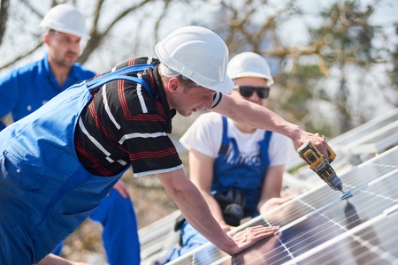 Male team workers installing stand-alone solar photovoltaic panel system using screwdriver. Electricians mounting blue solar module on roof of modern house. Alternative energy ecological concept.の写真素材