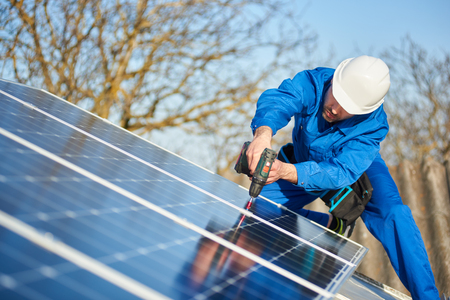 Male engineer in blue suit and protective helmet installing stand-alone solar photovoltaic panel system using screwdriver. Professional electrician mounting blue solar module on roof of modern houseの写真素材