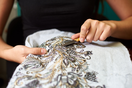 Close-up of female worker hands cutting away short loose ends of threads from white fabric clothing detail with machine embroidery of black and yellow floral pattern using special sharp scissors.の写真素材
