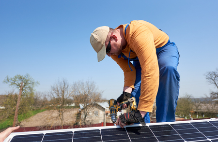 Male engineer installing stand-alone solar photovoltaic panel system using screwdriver. Electrician mounting blue solar module on roof of modern house. Alternative energy ecological concept.の写真素材
