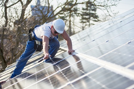 Male worker installing solar photovoltaic panel system using screwdriver. Electrician mounting blue solar module on roof of modern house. Alternative energy ecological concept.の写真素材