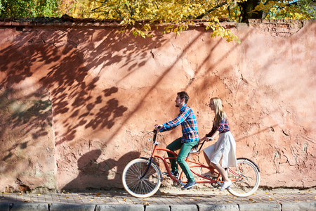 Young attractive happy tourist couple in casual clothing, bearded man and blond woman in glasses cycling together on double tandem bike on background of high red old crackled plastered wall.の写真素材