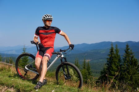 Young athletic tourist biker resting at his bike on mountain hill on bright blue summer sky and distant mountains background. Tourism, active lifestyle and extreme sport concept.の写真素材