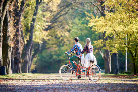 Back view of young romantic couple, man and blond woman cycling red tandem bike along autumn park or forest alley lit by bright sun and covered with golden leaves on green and yellow trees background.の写真素材