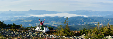 Panoramic view of camping on the top of mountain on bright summer morning. Back view of camper woman in pink sweater and sleeping bag standing in the entrance of tourist tent, holding hands lifting upの写真素材