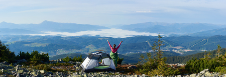 Panoramic view of camping on the top of mountain on summer morning. Back view of young hiker woman having a rest beside tourist tent, holding hands lifting up. On beautiful foggy mountains background.の写真素材