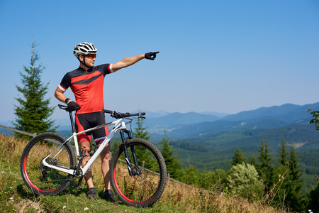 Young athletic professional biker standing with bicycle on top of hill, pointing into distance, enjoying view mountains, blue summer sky on background. Copy space. Active lifestyle and sport conceptの写真素材