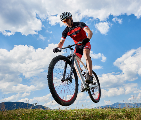 Young athletic sportsman biker in professional sportswear flying in air on his bike on bright blue sky with white clouds and distant hills background Active lifestyle and extreme sport concept.の写真素材