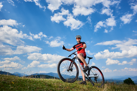 Smiling professional cyclist in sportswear and helmet standing with cross country bike on top of hill, showing thumbs up, against blue sky with clouds on summer sunny day. Outdoor sport conceptの写真素材