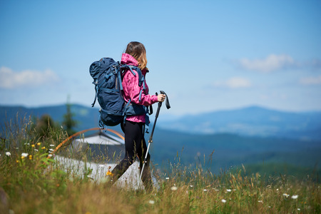 Back view of sporty girl traveller with backpack and trekking sticks near tent, wearing sports wear, hiking against blue sky and, enjoying sunny morning in the mountains. Camping lifestyle conceptの写真素材