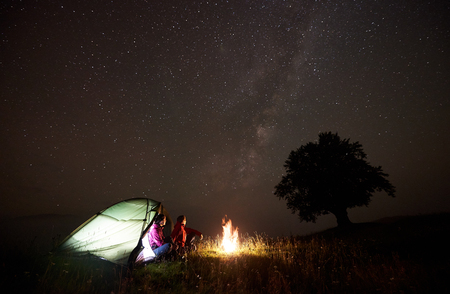 Tourist couple man and woman having a rest near illuminated tent, burning campfire and silhouette of big tree under starry sky, enjoying night camping in mountains. Tourism, outdoor activity conceptの写真素材