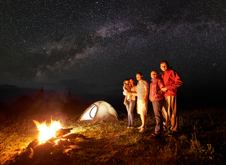 Night camping in mountains. Tourists standing in front of burning bonfire. Illuminated tent and starry sky on background. Woman holding in arms small daughter. Tourism and recreation concept.の写真素材