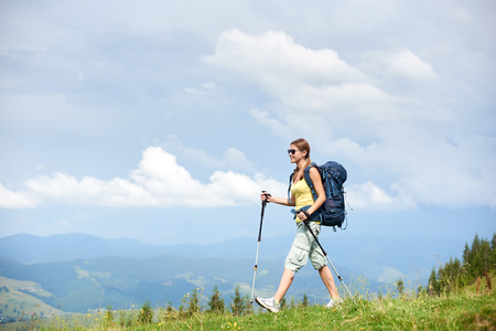 Smiling woman hiker hiking in Carpathian mountain trail, walking on grassy hill, wearing backpack and sunglasses, using trekking sticks, enjoying summer cloudy day. Outdoor activity, tourism conceptの写真素材