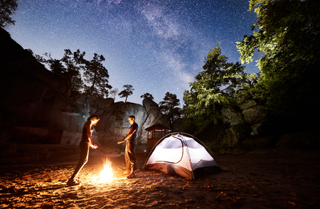Young couple tourists man and woman resting beside camp and illuminated tent at night, warming hands by campfire. On background night starry sky full of stars and Milky way, mountain rocks, treesの写真素材