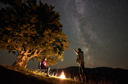 Romantic couple hikers having a rest at summer night camping in the mountains. Young woman sitting on chair beside campfire and big tree. Man pointing at amazing starry sky full of stars and Milky wayの写真素材