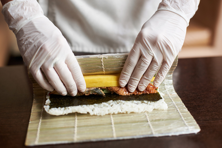 Master is making a sushi roll with nori, rice, cucumber, eel and omelet using bamboo mat. Closeup view of process of cooking sushiの写真素材