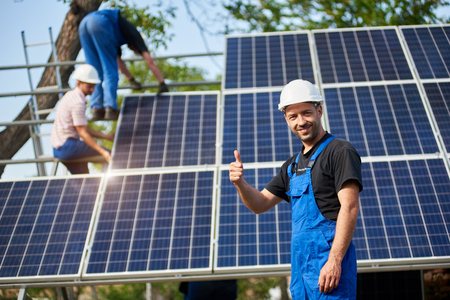 Portrait of happy technician showing thumb up gesture standing in front of unfinished high exterior solar panel photo voltaic system blue shiny surface with team of workers on high platform.の写真素材