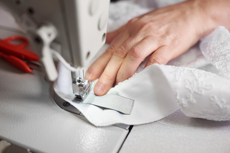 Close up front view of female hands sewing on auxiliary detail to white dress on professional sewing...