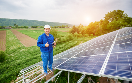 Construction worker with screwdriver looking in camera with thumb-up gesture on photo voltaic panel solar system shiny surface and lit by sun green fields background. Alternative energy concept.の写真素材