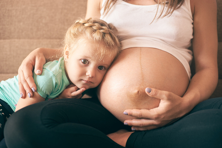 Close-up view of lovely girl sitting on sofa with pregnant mother and looking at camera. Cheerful woman embracing daughter and expecting baby. Concept of parental love and pregnancy.の写真素材