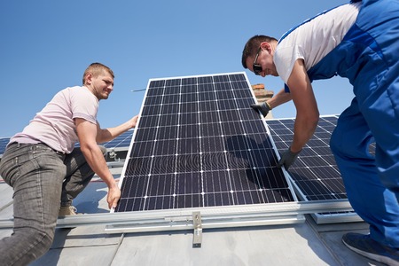 Male workers installing stand-alone solar photovoltaic panel system. Two electricians mounting blue solar module on roof of modern house. Alternative energy innovation ecology concept.の写真素材