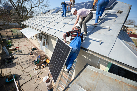 Male team workers installing stand-alone solar photovoltaic panel system. Electricians lifting blue solar module on roof of modern house. Alternative energy ecological concept.の写真素材
