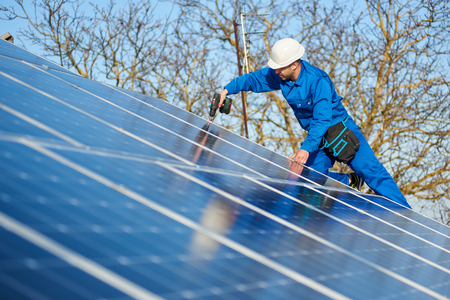 Man engineer in blue suit and protective helmet installing stand-alone solar photovoltaic panel system using screwdriver. Professional electrician mounting blue solar module on roof of modern house.