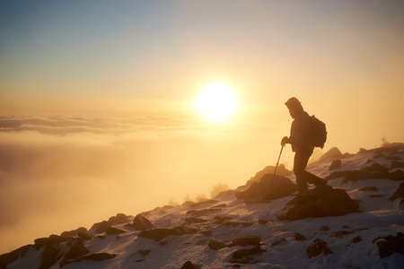 Silhouette of tourist hiker with backpack and trekking sticks hiking on rocky snowy mountain steep slope on background of foggy valley filled with white puffy clouds, raising sun and blue sky at dawn.の写真素材