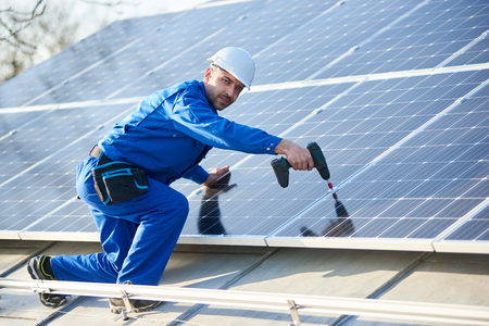 Man engineer in blue suit and helmet installing stand-alone solar photovoltaic panel system using screwdriver. Electrician mounting solar module on roof of modern house. Alternative energy concept.の写真素材