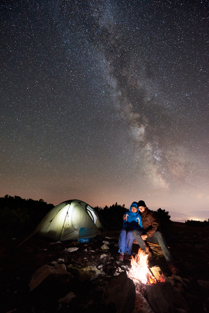 Happy couple hikers having a rest at night camping on the top of mountain. Young man hugging woman beside campfire, illuminated tourist tent under beautiful starry sky full of stars and Milky wayの写真素材