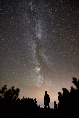 Silhouette of male hiker having a rest at summer night on the top of the mountain. Back view of man traveller enjoying view of night sky full of stars and Milky way.の写真素材